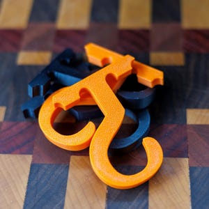 May include: An orange and black 3D-printed hook-shaped object, possibly a handle or a decorative element. The object is resting on a wooden surface with a checkered pattern of various wood tones. The hook has a textured surface.