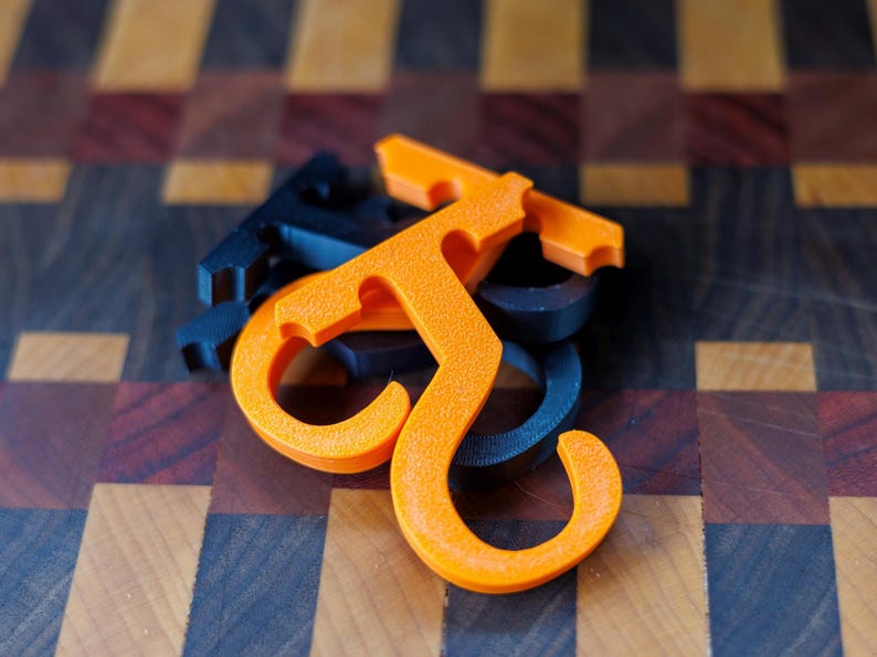 May include: An orange and black 3D-printed hook-shaped object, possibly a handle or a decorative element. The object is resting on a wooden surface with a checkered pattern of various wood tones. The hook has a textured surface.