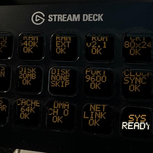 May include: A black Elgato Stream Deck controller featuring fifteen square keys with backlit orange text displays. The keys show various system status messages including CPU, RAM, DISK, PORT, CLOCK, BUS, CACHE, DMA, NET LINK, and SYS READY.
