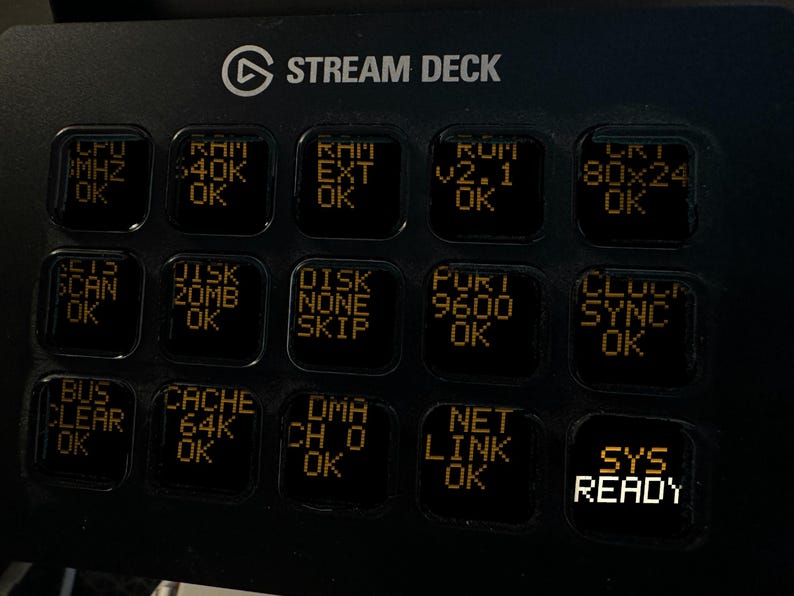 May include: A black Elgato Stream Deck controller featuring fifteen square keys with backlit orange text displays. The keys show various system status messages including CPU, RAM, DISK, PORT, CLOCK, BUS, CACHE, DMA, NET LINK, and SYS READY.