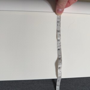 Pu&ograve; includere: Un oggetto rettangolare bianco viene misurato con un metro a nastro. Il metro a nastro mostra una misura di circa 18 cm. L'oggetto ha una parte superiore curva e si trova su una superficie scura.