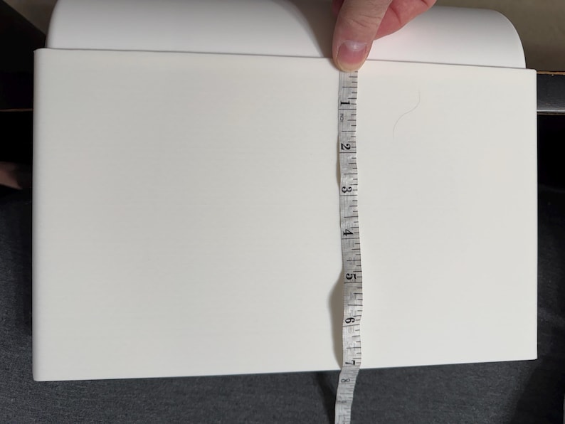 Pu&ograve; includere: Un oggetto rettangolare bianco viene misurato con un metro a nastro. Il metro a nastro mostra una misura di circa 18 cm. L'oggetto ha una parte superiore curva e si trova su una superficie scura.
