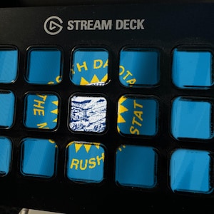 May include: A black Stream Deck controller featuring a grid of fifteen square buttons. Several buttons display segments of the blue and yellow North Dakota state flag, while one central button shows a detailed black and white landscape illustration.