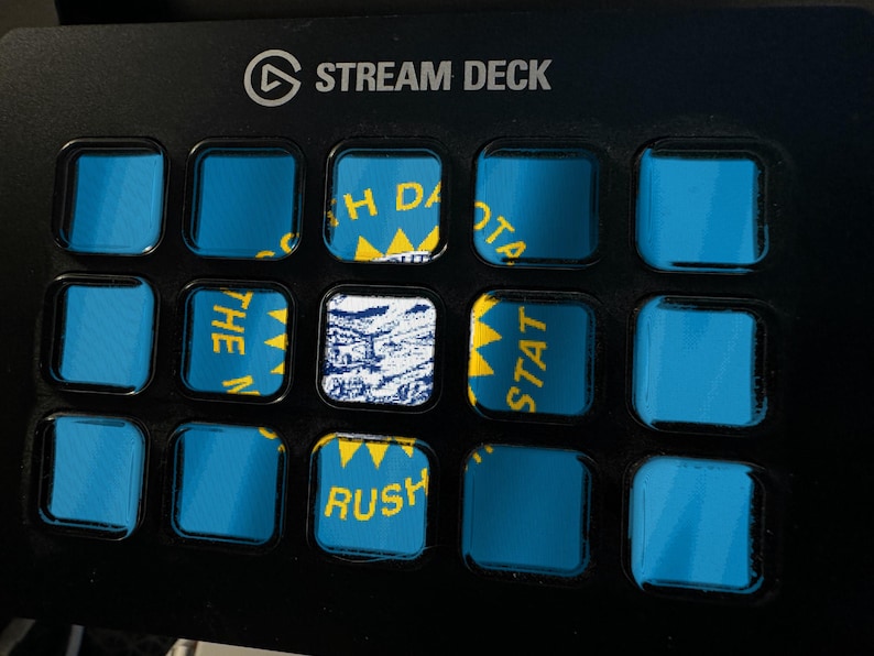 May include: A black Stream Deck controller featuring a grid of fifteen square buttons. Several buttons display segments of the blue and yellow North Dakota state flag, while one central button shows a detailed black and white landscape illustration.