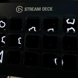 以下が含まれることがあります： 15個の正方形ボタンがグリッド状に配置された黒いStream Deckコントロールパネル。いくつかのボタンには、抽象的な図形やマウスカーソルなどの白い線画アイコンが暗い背景に照らされて表示されています。