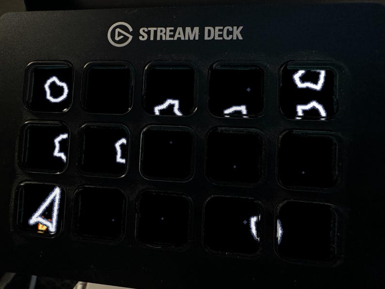 以下が含まれることがあります： 15個の正方形ボタンがグリッド状に配置された黒いStream Deckコントロールパネル。いくつかのボタンには、抽象的な図形やマウスカーソルなどの白い線画アイコンが暗い背景に照らされて表示されています。