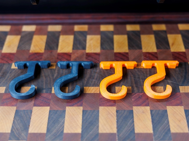 May include: Four T-shaped hooks, two in a dark blue-grey and two in bright orange, are arranged in a row. The hooks are made of a solid material and have a curved, J-shaped bottom. The hooks are displayed on a wooden surface with a checkered pattern.