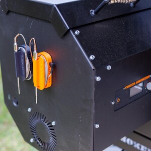 May include: Black smoker with an orange and black thermometer holder. The smoker has a digital display and a control knob. The brand name "Yoder Smokers" is visible on the front.