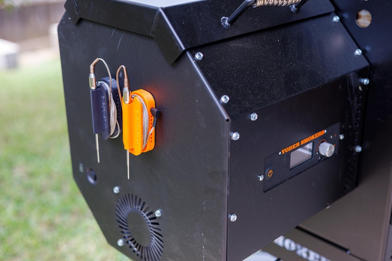 May include: Black smoker with an orange and black thermometer holder. The smoker has a digital display and a control knob. The brand name "Yoder Smokers" is visible on the front.