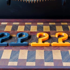 May include: Four 3D-printed, hook-shaped items. Two are dark blue, and two are bright orange. They are arranged on a wooden cutting board with a checkered pattern of various wood tones. The items have a flat base with text.