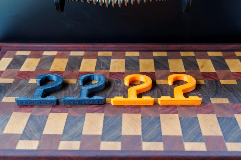 May include: Four 3D-printed, hook-shaped items. Two are dark blue, and two are bright orange. They are arranged on a wooden cutting board with a checkered pattern of various wood tones. The items have a flat base with text.