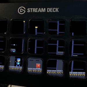 K&ouml;nnte beinhalten: Ein schwarzer Stream Deck Controller mit f&uuml;nfzehn quadratischen LCD-Tasten. Die Tasten zeigen eine verpixelte Videospielszene mit einer blauen Figur und einem roten Gegner auf einer Plattform mit gelb-schwarzen Warnstreifen. Der Text STREAM DECK ist sichtbar.
