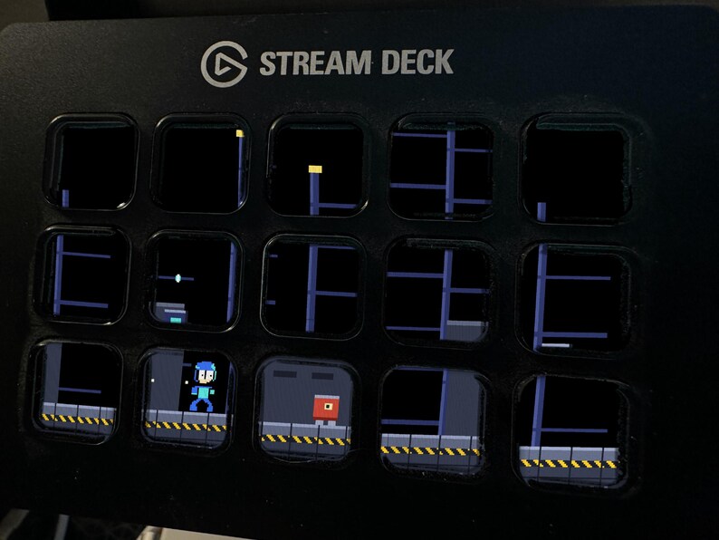 K&ouml;nnte beinhalten: Ein schwarzer Stream Deck Controller mit f&uuml;nfzehn quadratischen LCD-Tasten. Die Tasten zeigen eine verpixelte Videospielszene mit einer blauen Figur und einem roten Gegner auf einer Plattform mit gelb-schwarzen Warnstreifen. Der Text STREAM DECK ist sichtbar.