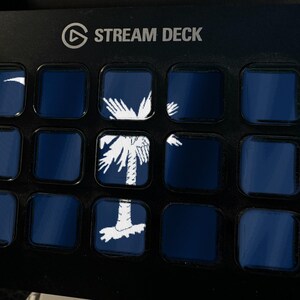 May include: A black Stream Deck control console featuring a grid of fifteen square buttons. Several buttons display a white graphic of a palmetto tree and crescent moon on a dark blue background, forming a larger image across the display.