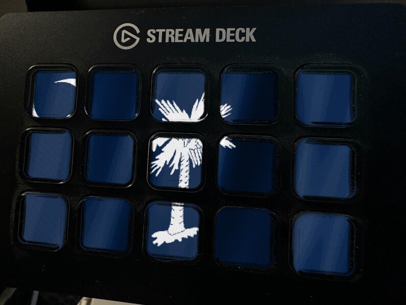 May include: A black Stream Deck control console featuring a grid of fifteen square buttons. Several buttons display a white graphic of a palmetto tree and crescent moon on a dark blue background, forming a larger image across the display.