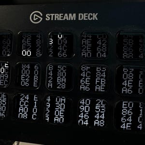 Puede incluir: Una consola de control Stream Deck negra con una cuadr&iacute;cula de quince teclas cuadradas. Cada tecla muestra una disposici&oacute;n &uacute;nica de caracteres alfanum&eacute;ricos blancos y c&oacute;digo hexadecimal, con algunos d&iacute;gitos iluminados m&aacute;s intensamente.