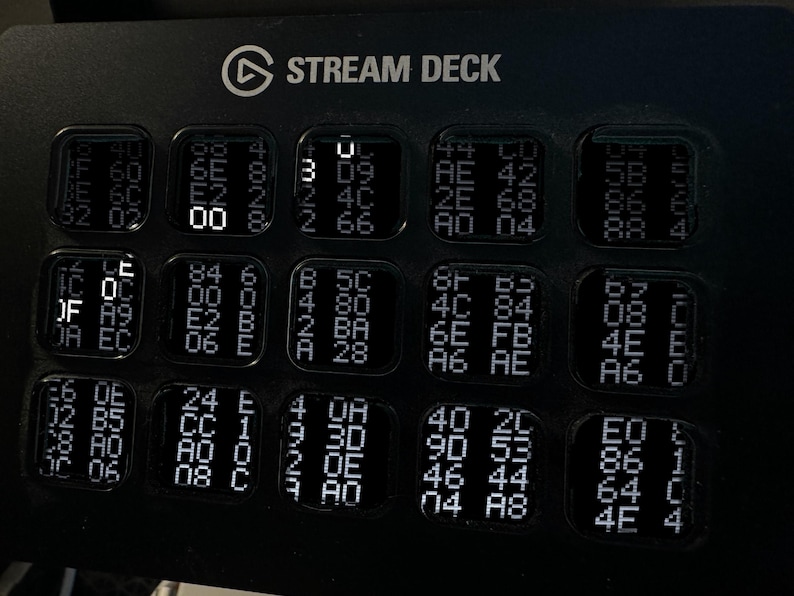 Puede incluir: Una consola de control Stream Deck negra con una cuadr&iacute;cula de quince teclas cuadradas. Cada tecla muestra una disposici&oacute;n &uacute;nica de caracteres alfanum&eacute;ricos blancos y c&oacute;digo hexadecimal, con algunos d&iacute;gitos iluminados m&aacute;s intensamente.