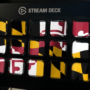 K&ouml;nnte beinhalten: Ein schwarzer Stream Deck-Controller mit f&uuml;nfzehn quadratischen Tasten in einem Raster. Jede Taste zeigt einen Teil der Flagge des Bundesstaates Maryland, die sich durch ihre markanten schwarzen, goldenen, wei&szlig;en und roten geometrischen Muster auszeichnet.
