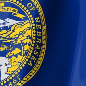 May include: A deep blue flag featuring the state seal of Nebraska in yellow. The seal displays a mountain landscape, a steamboat on a river, and a train, with a banner reading THE LAW and the text GREAT SEAL OF THE STATE OF NEBRASKA 1867.