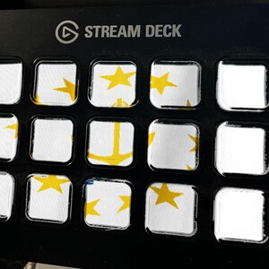 May include: A black Stream Deck controller featuring a grid of fifteen square keys. The keys display a white background with yellow star and anchor graphics, creating a fragmented image across the display surface of the device.