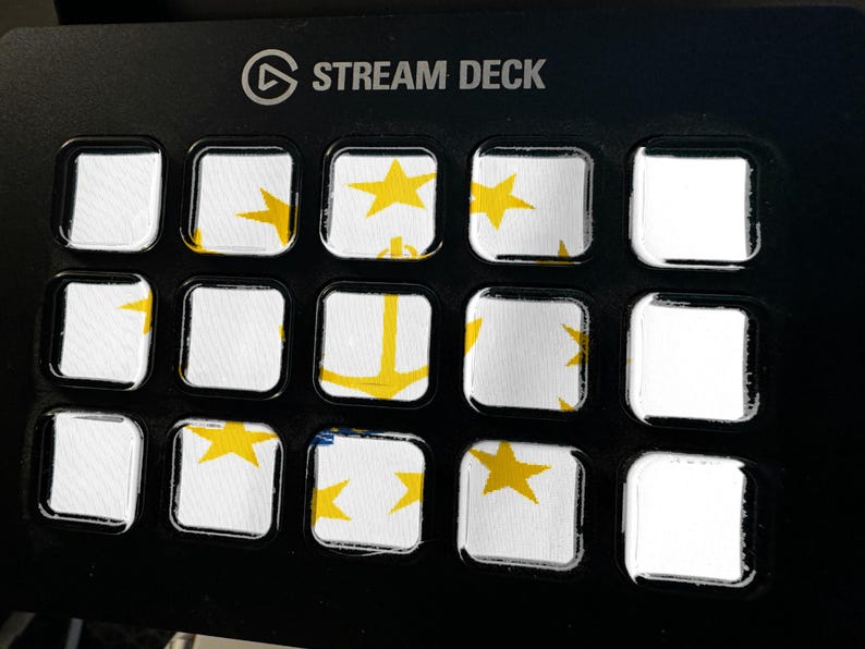May include: A black Stream Deck controller featuring a grid of fifteen square keys. The keys display a white background with yellow star and anchor graphics, creating a fragmented image across the display surface of the device.