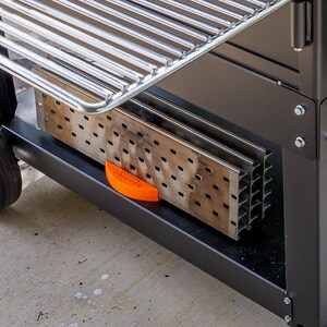 Op de afbeelding: Een zwarte buitengrill met een roestvrijstalen kookrooster. De grill heeft een onderste plank met een geperforeerde metalen inzet en een oranje veiligheidsvoorziening. Een zwart wiel is zichtbaar aan de linkerkant.