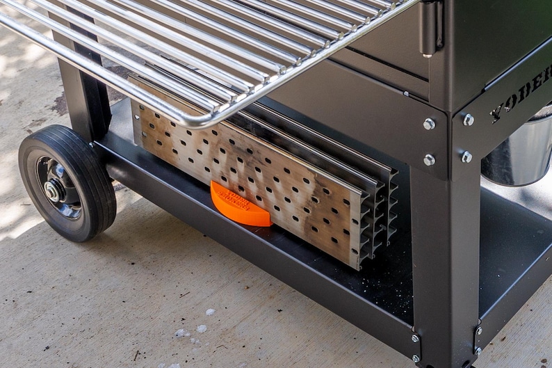 Op de afbeelding: Een zwarte buitengrill met een roestvrijstalen kookrooster. De grill heeft een onderste plank met een geperforeerde metalen inzet en een oranje veiligheidsvoorziening. Een zwart wiel is zichtbaar aan de linkerkant.