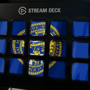 May include: A black Stream Deck device featuring a grid of fifteen square buttons. Several buttons display segments of the blue and yellow Nebraska state flag, including the state seal with text, while other buttons remain solid blue.