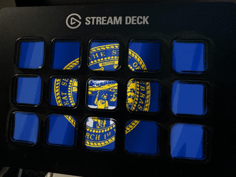May include: A black Stream Deck device featuring a grid of fifteen square buttons. Several buttons display segments of the blue and yellow Nebraska state flag, including the state seal with text, while other buttons remain solid blue.