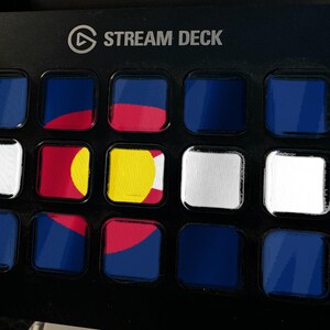 Pode incluir: Um controlador Stream Deck preto com uma grade de quinze bot&otilde;es quadrados. V&aacute;rios bot&otilde;es exibem segmentos da bandeira do estado do Colorado, com cores azul, vermelho, amarelo e branco, enquanto outros s&atilde;o de cor azul ou branca s&oacute;lida.