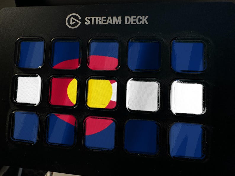 Pode incluir: Um controlador Stream Deck preto com uma grade de quinze bot&otilde;es quadrados. V&aacute;rios bot&otilde;es exibem segmentos da bandeira do estado do Colorado, com cores azul, vermelho, amarelo e branco, enquanto outros s&atilde;o de cor azul ou branca s&oacute;lida.