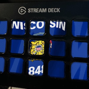 Peut inclure: Un contr&ocirc;leur Stream Deck noir dot&eacute; d'une grille de quinze boutons carr&eacute;s. Plusieurs boutons affichent des graphiques partiels bleus et blancs, dont le texte 'WISCONSIN', un sceau d'&Eacute;tat et le nombre '848', tandis que d'autres restent bleu uni.