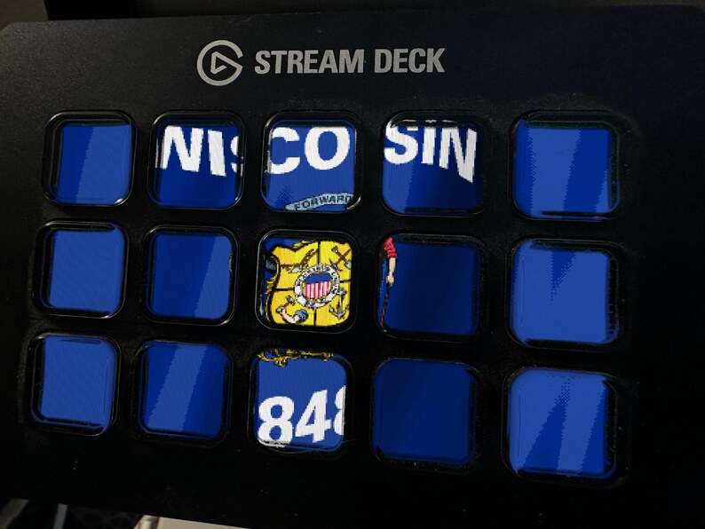 Peut inclure: Un contr&ocirc;leur Stream Deck noir dot&eacute; d'une grille de quinze boutons carr&eacute;s. Plusieurs boutons affichent des graphiques partiels bleus et blancs, dont le texte 'WISCONSIN', un sceau d'&Eacute;tat et le nombre '848', tandis que d'autres restent bleu uni.