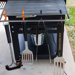 May include: A black grill with a stainless steel grate and a set of grilling tools hanging below. The tools include a spatula, tongs, and a brush. The grill has the words "POWER SMOKERS" and "GRILLGRATE" printed on it.