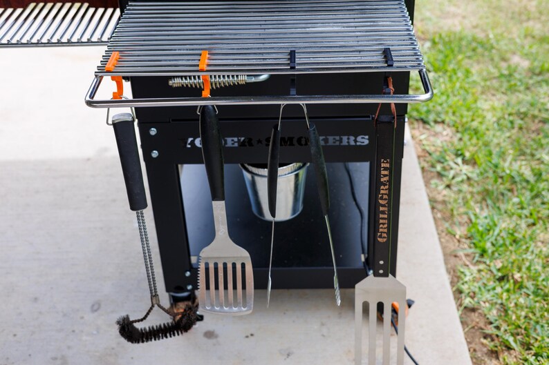 May include: A black grill with a stainless steel grate and a set of grilling tools hanging below. The tools include a spatula, tongs, and a brush. The grill has the words "POWER SMOKERS" and "GRILLGRATE" printed on it.