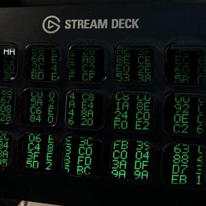 Op de afbeelding: Een zwarte Elgato Stream Deck-controller met vijftien vierkante LCD-toetsen in een raster. Elke toets toont een uniek patroon van groene alfanumerieke tekens en hexadecimale codes tegen een donkere achtergrond.