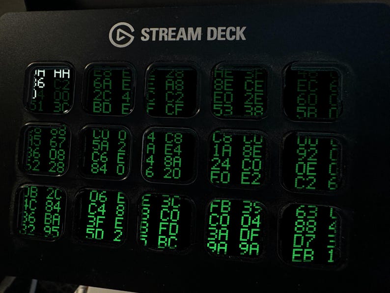 Op de afbeelding: Een zwarte Elgato Stream Deck-controller met vijftien vierkante LCD-toetsen in een raster. Elke toets toont een uniek patroon van groene alfanumerieke tekens en hexadecimale codes tegen een donkere achtergrond.