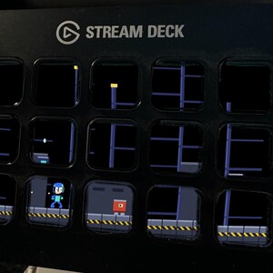 May include: A black Stream Deck control console featuring fifteen square LCD keys. The keys display a pixelated side-scrolling video game interface with a blue character, a red enemy, and industrial platform levels with yellow hazard stripes.
