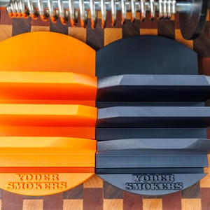 Op de afbeelding: Twee Yoder Smokers-schapen, &eacute;&eacute;n oranje en &eacute;&eacute;n donkergrijs, worden getoond. Elk schap heeft drie niveaus en een halfronde basis. De schappen staan tegen een achtergrond van een houten snijplank.