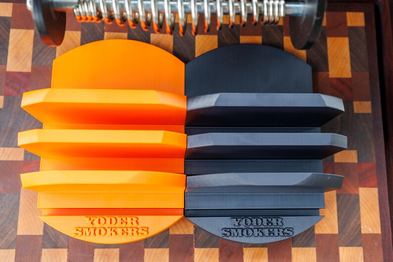 Op de afbeelding: Twee Yoder Smokers-schapen, &eacute;&eacute;n oranje en &eacute;&eacute;n donkergrijs, worden getoond. Elk schap heeft drie niveaus en een halfronde basis. De schappen staan tegen een achtergrond van een houten snijplank.