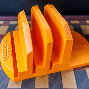 Op de afbeelding: Fel oranje sandwichhouder met drie verticale sleuven. De basis is halfrond met de tekst "KODER SANDWICHES" in reli&euml;f aan de zijkant. De houder is gemaakt van een solide materiaal en staat op een houten snijplank.