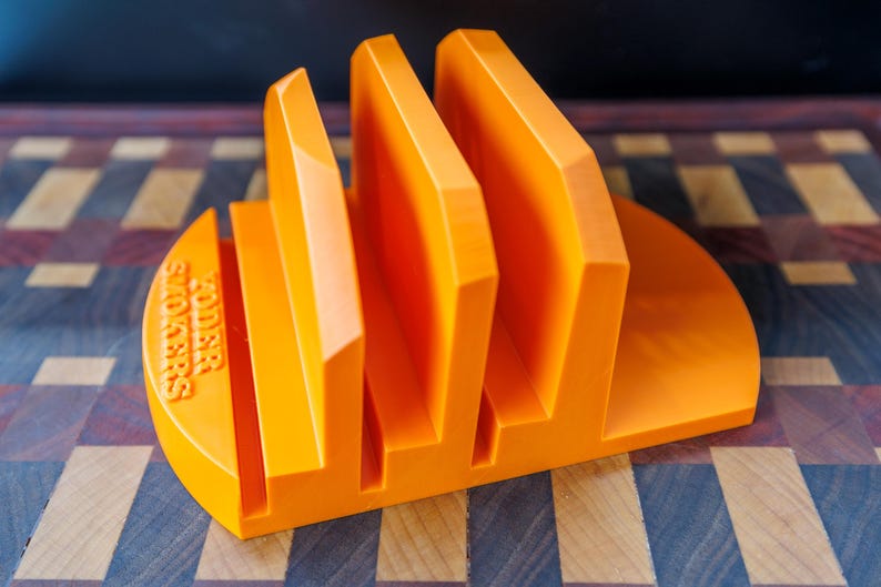 Op de afbeelding: Fel oranje sandwichhouder met drie verticale sleuven. De basis is halfrond met de tekst "KODER SANDWICHES" in reli&euml;f aan de zijkant. De houder is gemaakt van een solide materiaal en staat op een houten snijplank.