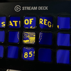 Może przedstawiać: Czarne urządzenie Stream Deck z siatką piętnastu kwadratowych klawiszy LCD. Klawisze wyświetlają fragmenty flagi stanu Oregon w kolorze niebieskim i ż&oacute;łtym, w tym tekst 'STATE OF OREGON', ilustrację wozu osadnik&oacute;w oraz liczbę '1859'.