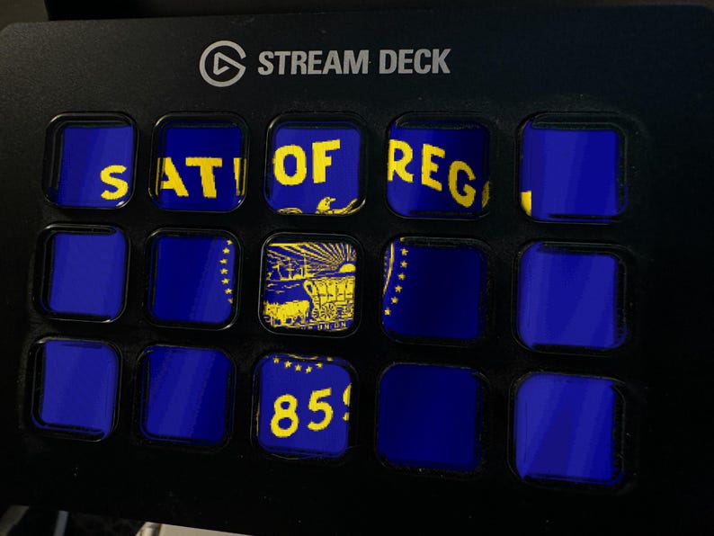 Może przedstawiać: Czarne urządzenie Stream Deck z siatką piętnastu kwadratowych klawiszy LCD. Klawisze wyświetlają fragmenty flagi stanu Oregon w kolorze niebieskim i ż&oacute;łtym, w tym tekst 'STATE OF OREGON', ilustrację wozu osadnik&oacute;w oraz liczbę '1859'.