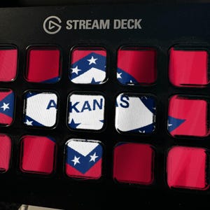 May include: A black Stream Deck controller featuring a grid of fifteen square buttons. Several buttons display segments of the Arkansas state flag, including the white diamond, blue border, and red background, with the text ARKANSAS visible.