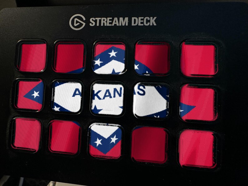 May include: A black Stream Deck controller featuring a grid of fifteen square buttons. Several buttons display segments of the Arkansas state flag, including the white diamond, blue border, and red background, with the text ARKANSAS visible.