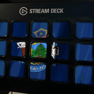 May include: A black Stream Deck control panel featuring fifteen square buttons arranged in a grid. Several buttons display segments of the Maine state flag, including a green pine tree, a moose, and the text MAINE, while others remain dark blue.
