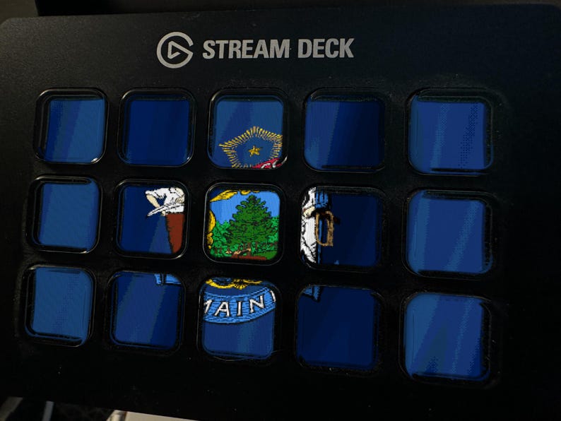 May include: A black Stream Deck control panel featuring fifteen square buttons arranged in a grid. Several buttons display segments of the Maine state flag, including a green pine tree, a moose, and the text MAINE, while others remain dark blue.