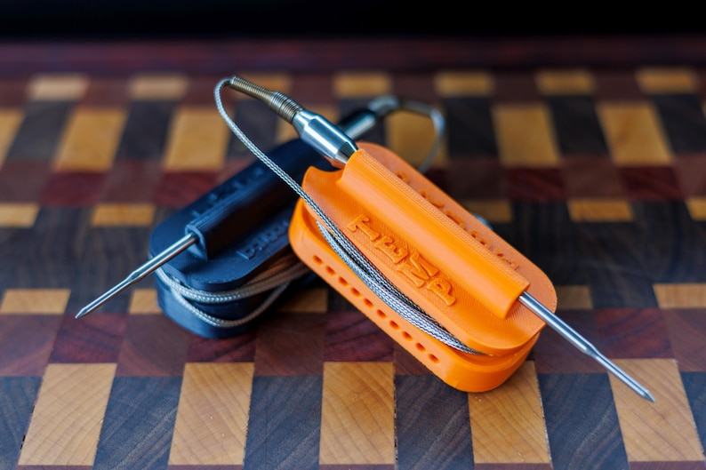 May include: Two food thermometers with stainless steel probes. One is orange and the other is black. The orange thermometer has the word "TEMP" printed on it. Both thermometers have a wire attached to the probe.