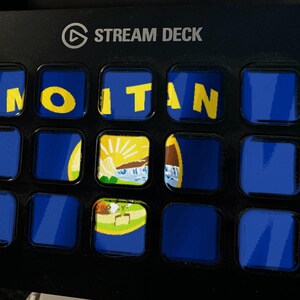 Puede incluir: Un controlador Stream Deck negro con una cuadr&iacute;cula de quince botones cuadrados. Los botones muestran un fondo azul con el texto amarillo MONTANA y un gr&aacute;fico parcial del sello estatal, con varios botones mostrando solo azul.